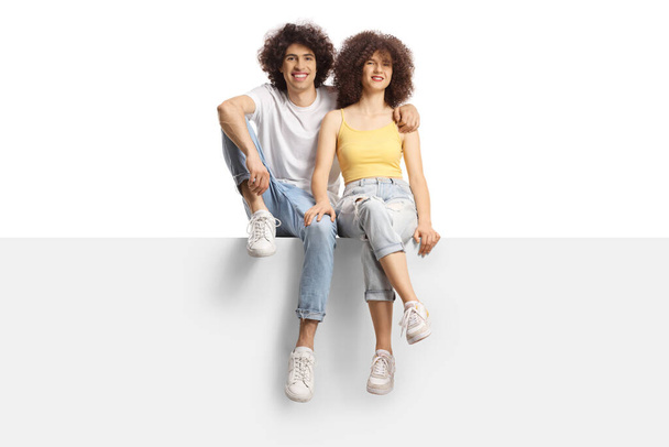 Young man and woman with curly hair smiling sitting on a blank panel isolated on white background - Photo, Image