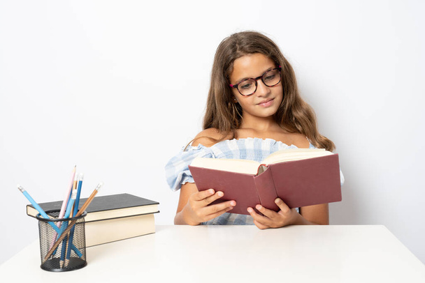 Портрет позитивной школьницы, сидящей за столом и читающей книгу на белом фоне - Фото, изображение