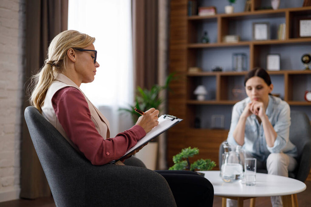 Upset young woman has counseling session with female psychologist in the office, telling her about problems. Psychologist taking notes on clipboard. Psychology, mental therapy, mental health. - Photo, image