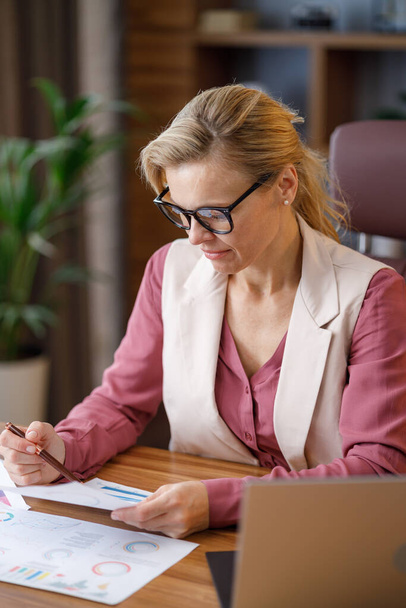 Portrait of adult businesswoman wearing eyeglasses working at office. Female manager doing paperwork, analyzing financial report, holding documents with graphs and charts at workplace. - Foto, immagini