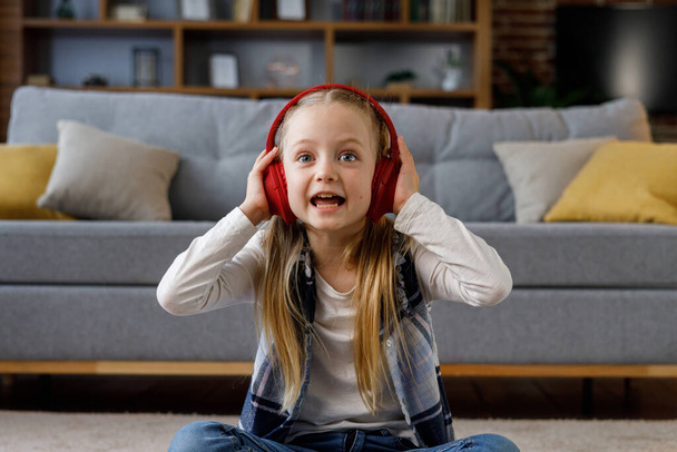 Little girl wearing red earphones looking at camera. Portrait of cute smiling child resting indoors at home interior. Concept of a happy childhood. - Photo, image