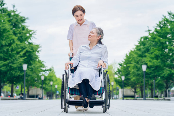 Жінка в інвалідному візку і молода жінка в фартусі для догляду на відкритому повітрі
 - Фото, зображення