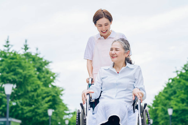 Una mujer en silla de ruedas y una mujer joven en un delantal para cuidar al aire libre - Foto, Imagen