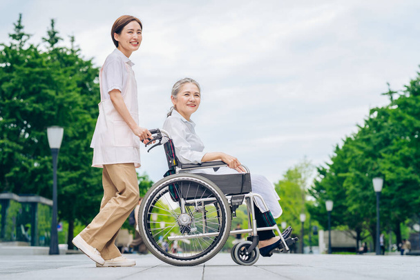 Eine Frau im Rollstuhl und eine junge Frau in einer Schürze, die sich um die Pflege im Freien kümmert - Foto, Bild