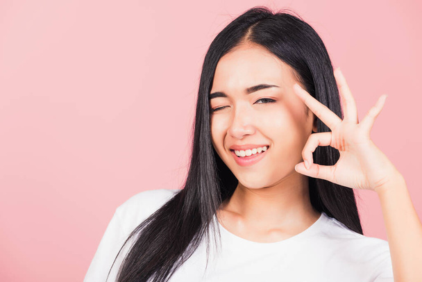 Portrait Asian beautiful young woman made finger OK symbol sign gesture wink eyes to agree and looking at camera, studio shot isolated on pink background, Thai female agree hand sign copy space - Foto, Imagen