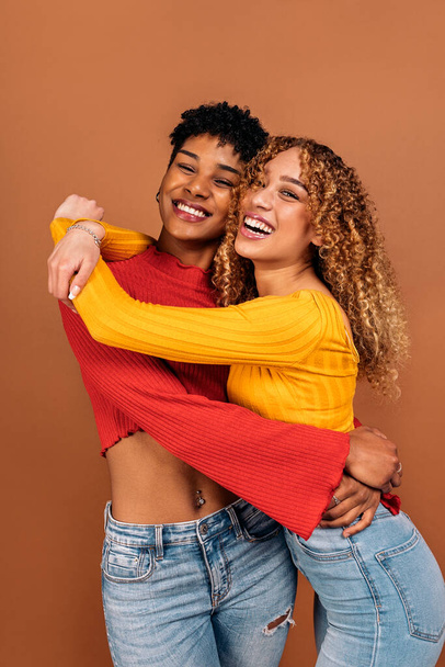 Stock photo of cheerful afro women hugging each other in studio shot against brown background. - Foto, imagen