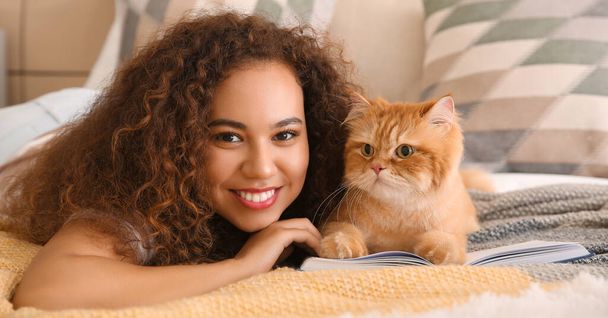 Young African-American woman with cute cat reading book in bedroom - Zdjęcie, obraz