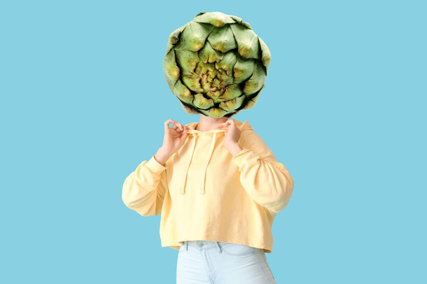 Young woman with fresh artichoke instead of her head on blue background - Fotó, kép