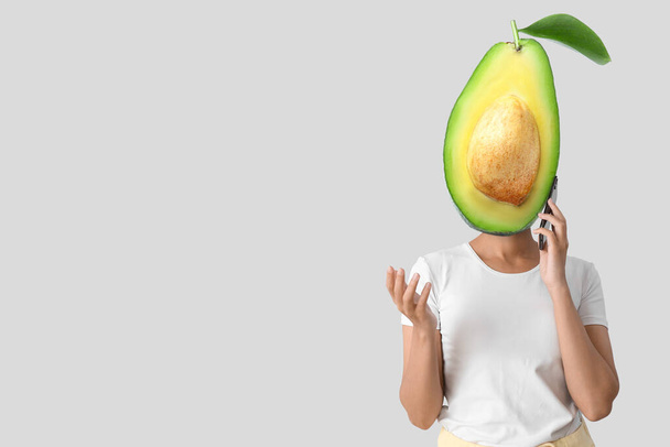 Young woman with fresh avocado instead of her head and mobile phone on grey background with space for text - Foto, imagen