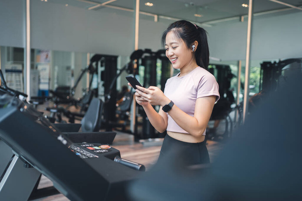 Woman exercising on treadmill wearing wireless earphones and smart watch checking mobile phone. - Photo, Image