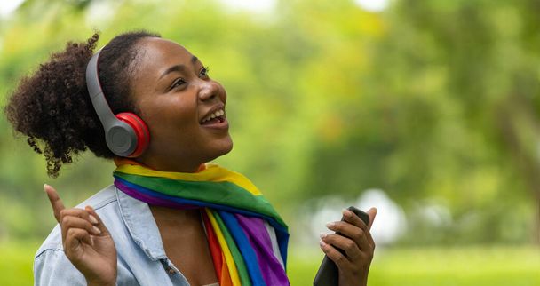 Africký Američan LGBTQ + žena poslouchá hudbu z jejího smartphonu, zatímco sedí relaxačně ve veřejném parku během léta - Fotografie, Obrázek