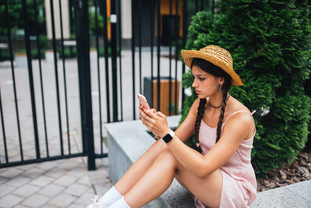 Jeune femme mince est assis sur un banc avec téléphone mobile, valise grise debout par les jambes, photo de côté - Photo, image