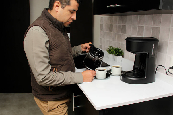 Homme adulte latino aux cheveux bruns prépare une tasse de café dans une cafetière pour sentir et goûter au petit déjeuner dans la cuisine est réfléchi, inquiet, réfléchi, déprimé - Photo, image
