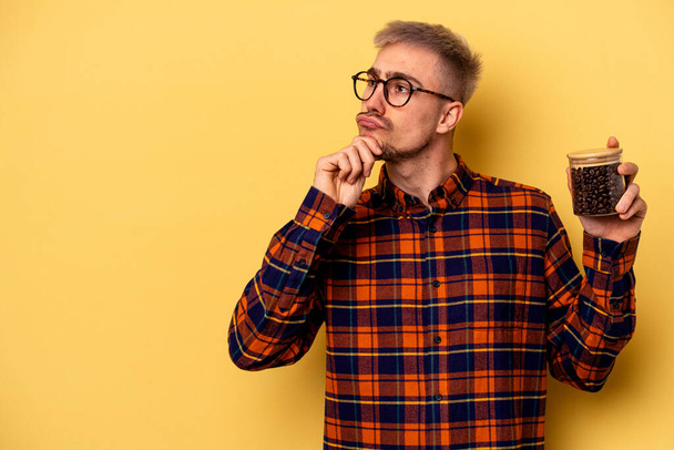 Young caucasian man holding coffee jar isolated on yellow background looking sideways with doubtful and skeptical expression. - Foto, afbeelding
