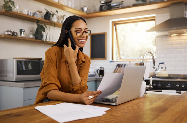 Young happy mixed race businesswoman going through paper and bills while on a call using her phone and working on a laptop at home. One hispanic female businessperson using a laptop and talking on a - Valokuva, kuva