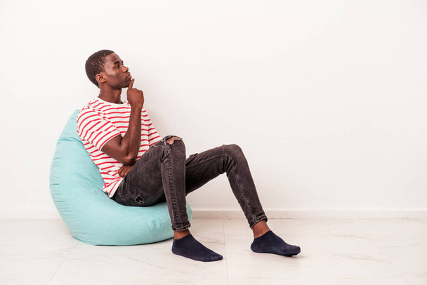 Jeune homme afro-américain assis sur une bouffée isolé sur fond blanc regardant de côté avec une expression douteuse et sceptique. - Photo, image