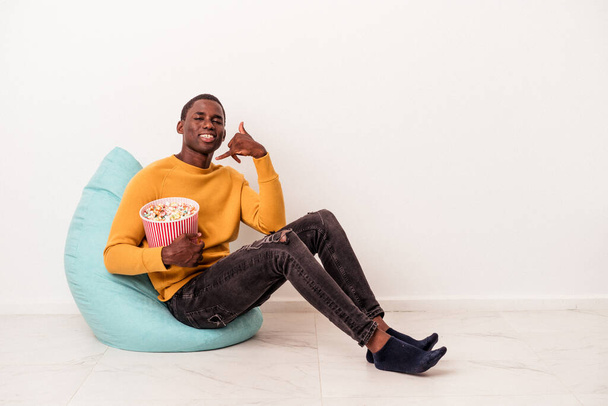 Junger afroamerikanischer Mann sitzt auf einem Puff und isst Popcorn isoliert auf weißem Hintergrund und zeigt eine Handygeste mit den Fingern. - Foto, Bild