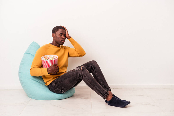 Jonge Afro-Amerikaanse man zit op een trekje eten popcorn geïsoleerd op witte achtergrond wordt geschokt, ze heeft onthouden belangrijke vergadering. - Foto, afbeelding