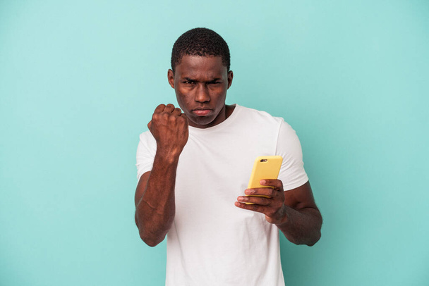 Elinde cep telefonu olan genç Afrikalı bir adam mavi arka planda izole edilmiş yumruğunu kameraya gösteriyor, agresif bir yüz ifadesi.. - Fotoğraf, Görsel