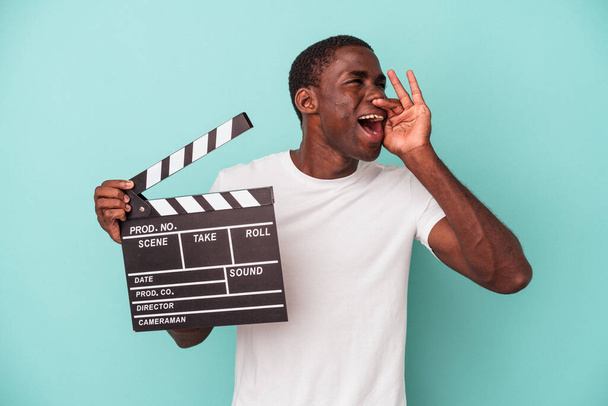młody Afroamerykanin trzyma klapperboard odizolowany na niebieskim tle krzycząc i trzymając dłoń w pobliżu otwartych ust. - Zdjęcie, obraz