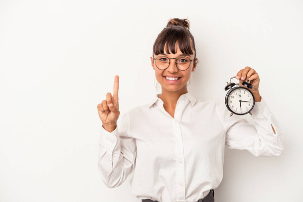 Joven mujer de negocios de raza mixta sosteniendo un reloj despertador aislado sobre fondo blanco mostrando el número uno con el dedo. - Foto, Imagen