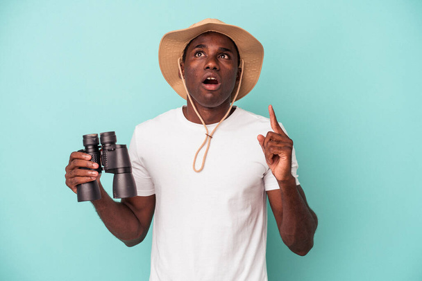 Joven afroamericano sosteniendo binoculares aislados sobre fondo azul apuntando al revés con la boca abierta. - Foto, imagen