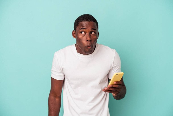 Young African American man holding a mobile phone isolated on blue background shrugs shoulders and open eyes confused. - Photo, image