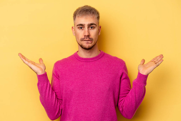 Young caucasian man isolated on yellow background confused and doubtful shrugging shoulders to hold a copy space. - Photo, Image