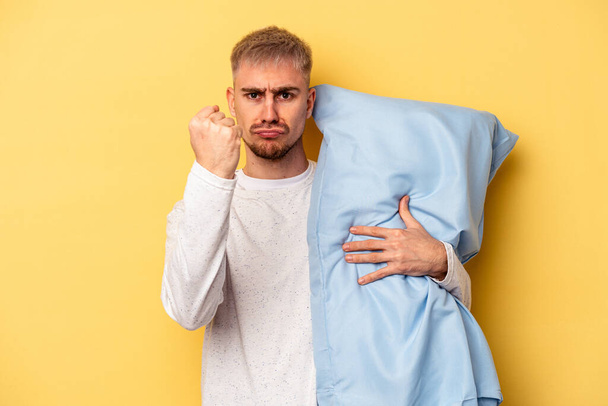 Young caucasian man wearing a pajama and holding pillow isolated on yellow background showing fist to camera, aggressive facial expression. - Fotó, kép