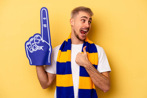 Young sports fan caucasian man isolated on yellow background points with thumb finger away, laughing and carefree. - Foto, immagini