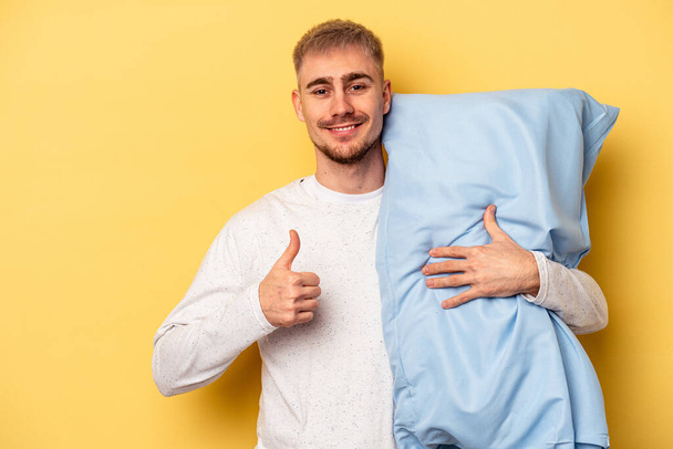 Young caucasian man wearing a pajama and holding pillow isolated on yellow background smiling and raising thumb up - Foto, imagen