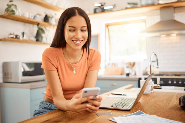 Young beautiful happy mixed race businesswoman holding and using a phone while working from home. One content hispanic woman using social media on her cellphone while using a laptop sitting at a - Фото, зображення