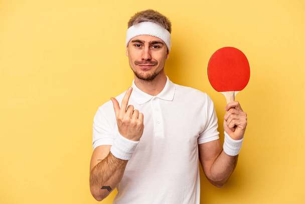 Молодой кавказский человек держит ракетки для пинг-понга изолированные на желтом фоне указывая пальцем на вас, как будто приглашая подойти ближе. - Фото, изображение
