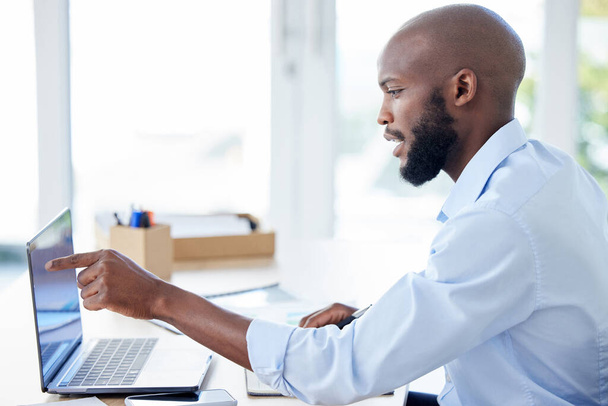 Jovem empresário afro-americano apontando o dedo e trabalhando em um laptop em um escritório sozinho no trabalho. Um homem de negócios usando um laptop em uma mesa no trabalho. - Foto, Imagem