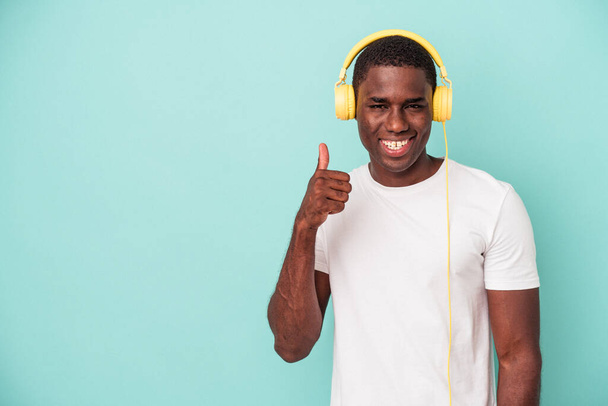 Jeune homme afro-américain à l'écoute de musique isolée sur fond bleu souriant et levant le pouce - Photo, image
