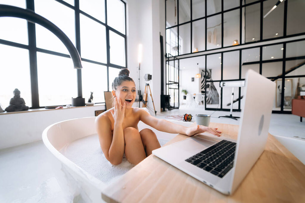 Jovem mulher trabalhando no laptop enquanto toma uma banheira em casa - Foto, Imagem
