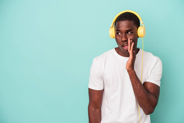 Młody, afrykański Amerykanin słuchający muzyki odizolowanej na niebieskim tle mówi sekretną wiadomość o gorącym hamowaniu i odwraca wzrok. - Zdjęcie, obraz