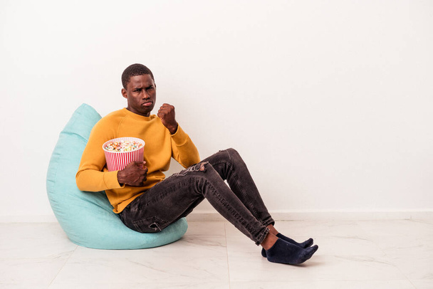 Junger afroamerikanischer Mann sitzt auf einem Puff und isst Popcorn isoliert auf weißem Hintergrund und zeigt der Kamera die Faust, aggressiver Gesichtsausdruck. - Foto, Bild