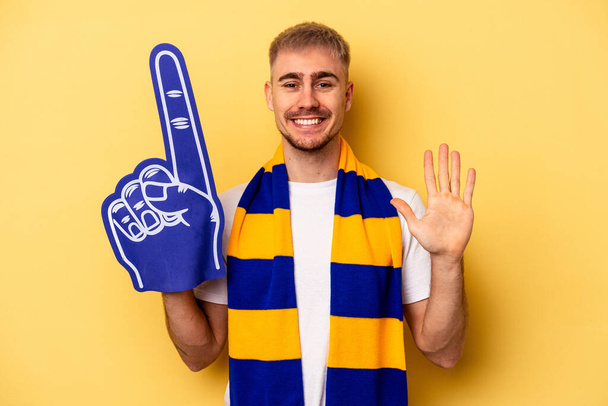 Young sports fan caucasian man isolated on yellow background smiling cheerful showing number five with fingers. - Zdjęcie, obraz