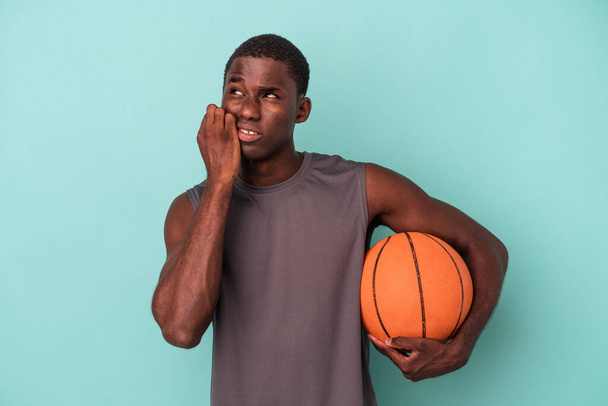若いですアフリカ系アメリカ人男性プレーバスケットボールの青の背景に隔離された爪をかむ、神経と非常に不安. - 写真・画像