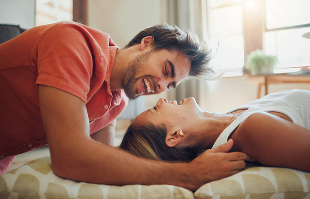 Happy young woman lying down while her boyfriend leans over her as they laugh together and look into each other eyes. Affectionate couple enjoying romantic intimate moment at home. - 写真・画像