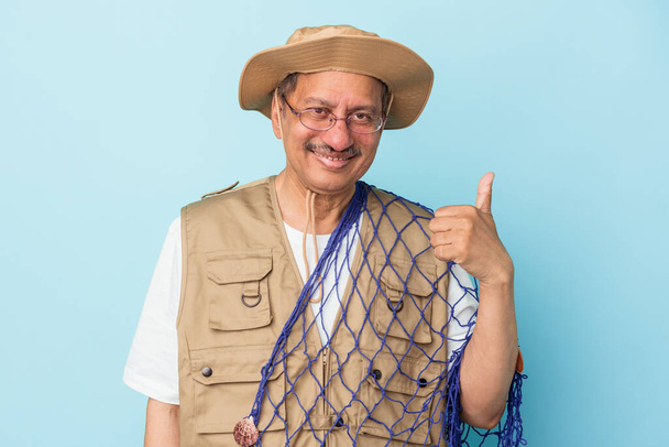 Senior indian fisherman holding net isolated on blue background smiling and raising thumb up - Foto, Imagen
