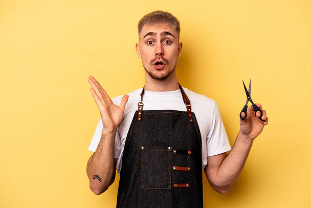 Young caucasian hairdresser man holding scissors isolated on yellow background surprised and shocked. - Foto, imagen