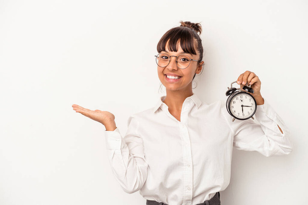 Young mixed race business woman holding an alarm clock isolated on white background  showing a copy space on a palm and holding another hand on waist. - Photo, Image