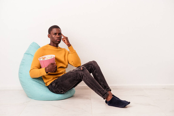 Junger afroamerikanischer Mann sitzt auf einem Puff und isst Popcorn isoliert auf weißem Hintergrund und zeigt mit dem Finger auf die Schläfe, denkt nach, konzentriert auf eine Aufgabe. - Foto, Bild