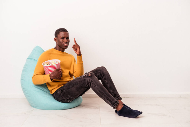 Jonge Afro-Amerikaanse man zit op een trekje popcorn te eten geïsoleerd op witte achtergrond toont nummer een met vinger. - Foto, afbeelding