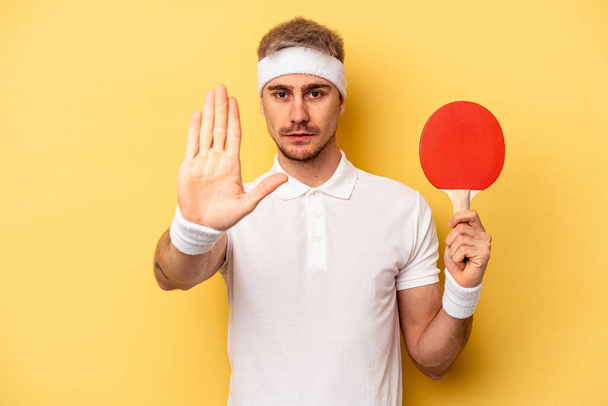 Young caucasian man holding ping pong rackets isolated on yellow background standing with outstretched hand showing stop sign, preventing you. - Φωτογραφία, εικόνα
