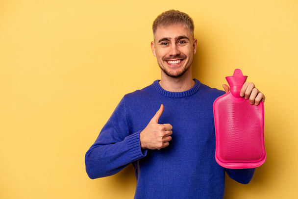 Young caucasian man holding a water bag isolated on yellow background smiling and raising thumb up - Фото, зображення