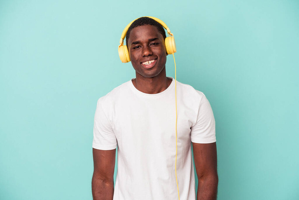 Młody, afrykański mężczyzna słuchający muzyki odizolowanej na niebieskim tle szczęśliwy, uśmiechnięty i pogodny. - Zdjęcie, obraz