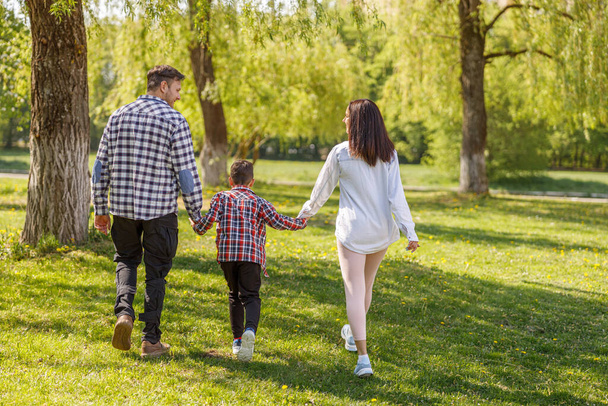Zadní na bělocha muž, žena a syn kráčející ruku v ruce v zeleném letním parku. Zadní pohled na rodinu. - Fotografie, Obrázek